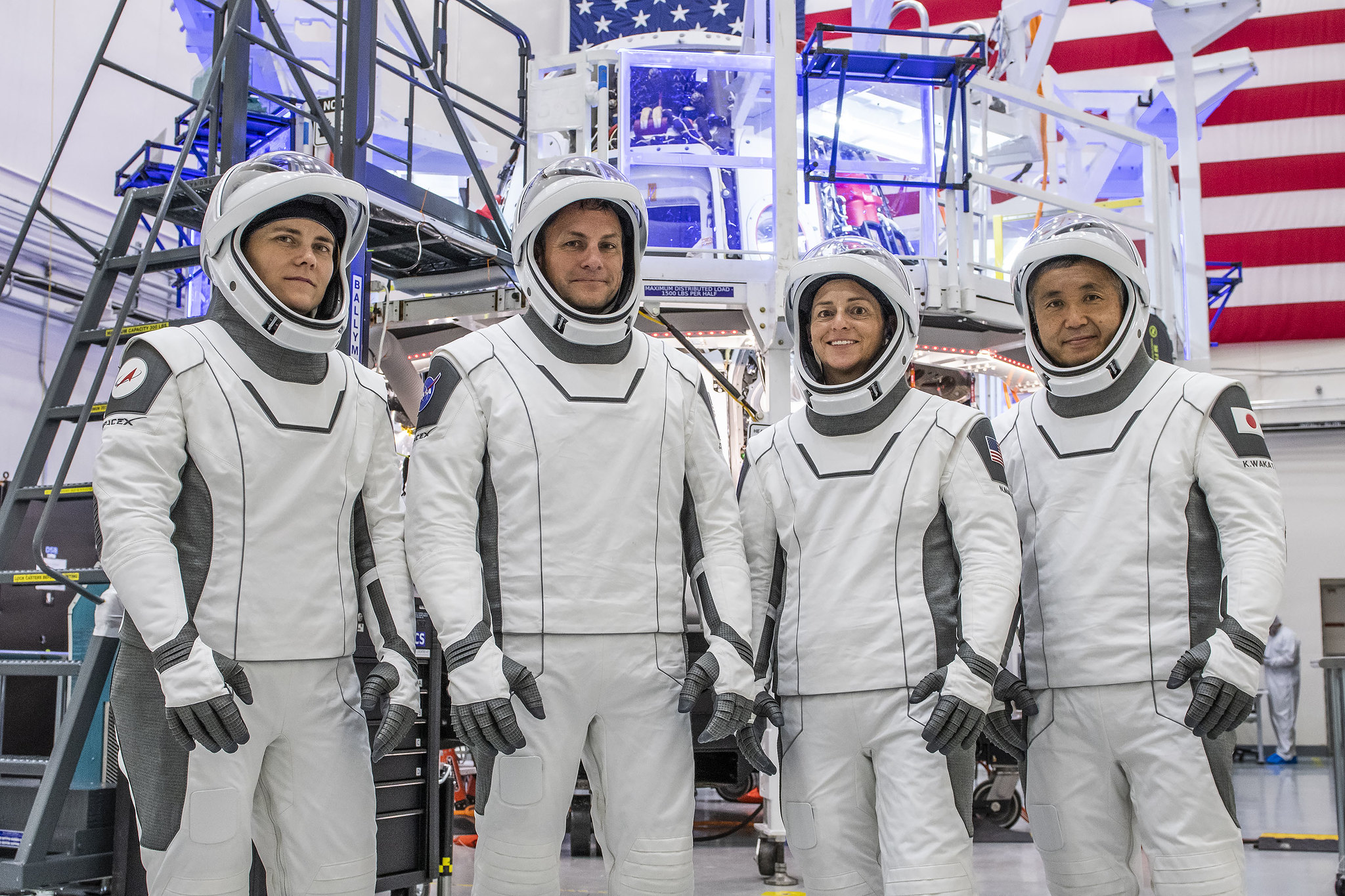 four astronauts in white spacesuits line up in front of space hardware