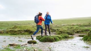 Two women jumping over a stream while out hiking, wearing hiking boots and hiking clothing, laughing and smiling after learning how to keep your feet warm