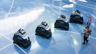 A photo of four automated floor scrubbers, robots created by Carnegie Robotics within Pittsburgh International Airport, while a woman with two young children walk past to the right of the frame. The robots have been pictured from above, and are sat on a blue polished floor.