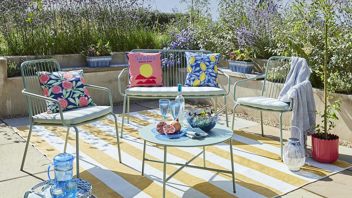 A blue outdoor furniture set with seat cushions on a garden terrace with raised beds full of summer planting in the background