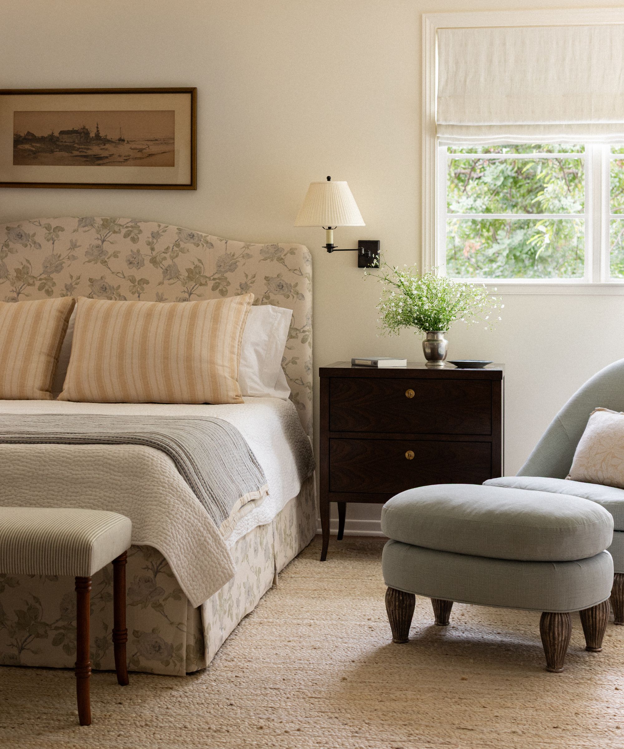 a neutral primary bedroom with a floral upholstered bed, foot of bed bench, black nightstand and a small armchair and ottoman