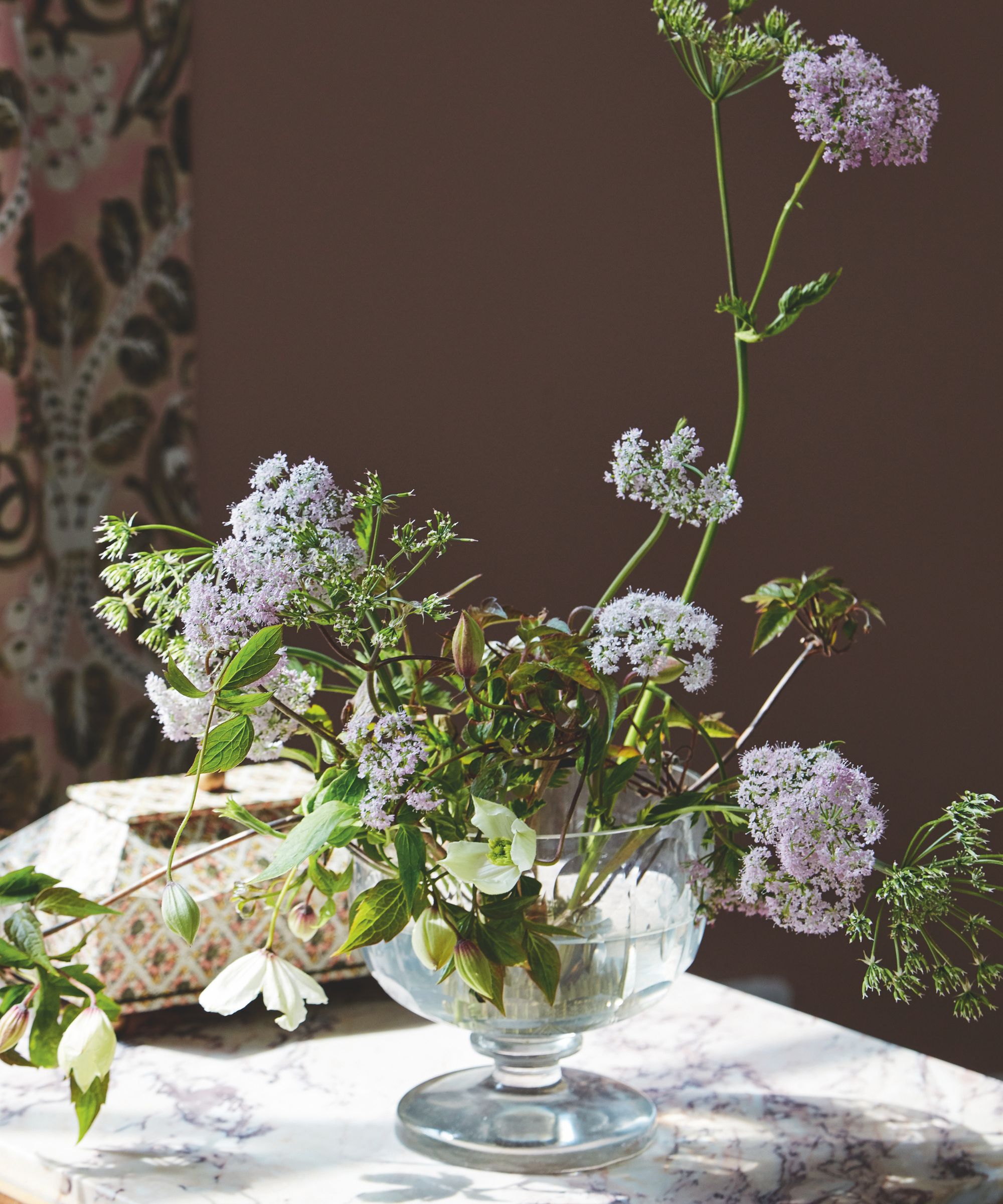 A glass vase of purple flowers against a brown wall on a marble surface.