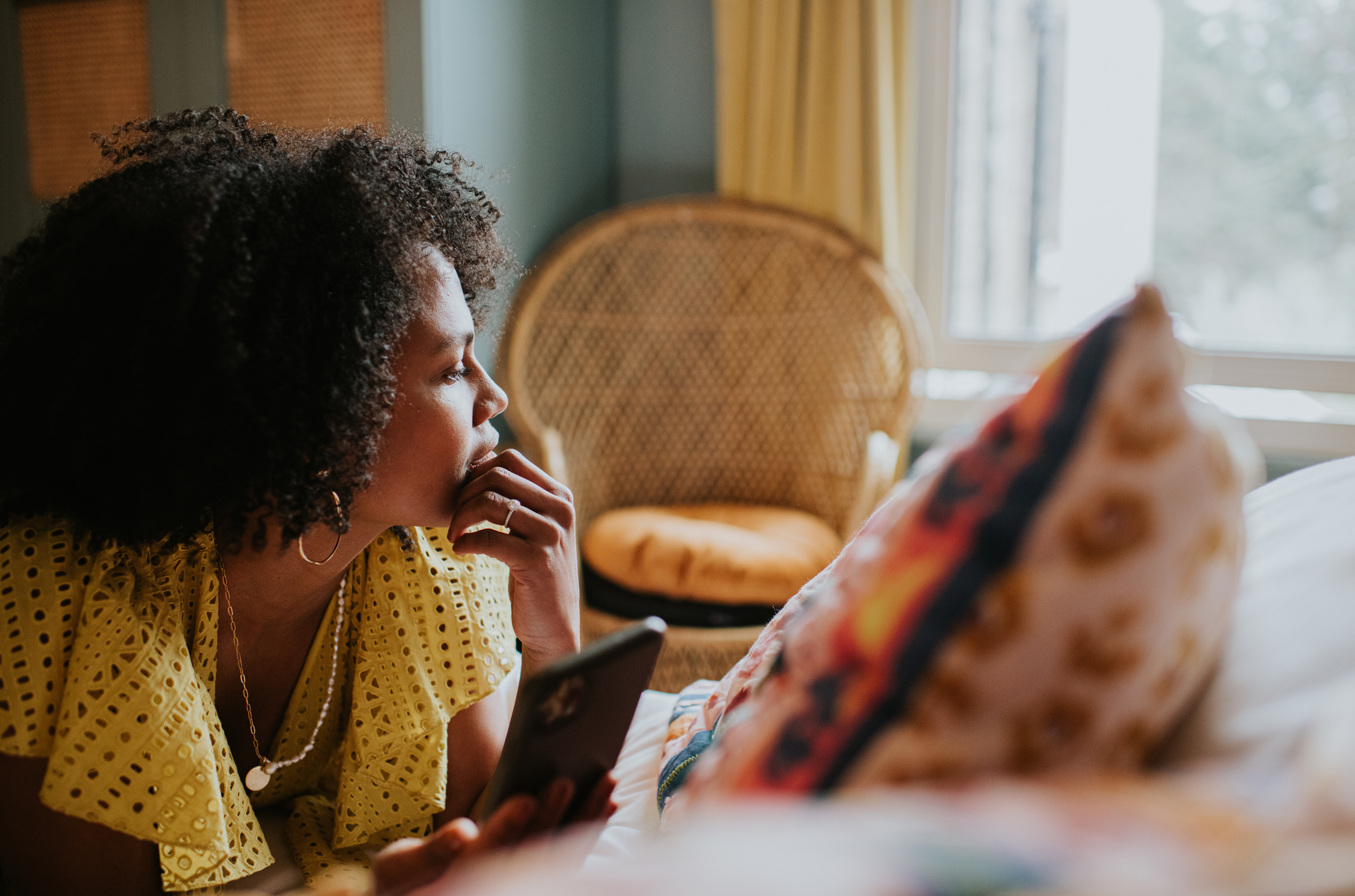 How to avoid Black Friday burnout – a woman looks distracted and pensive as she lies on a bed holding a mobile phone, and gazes out the window