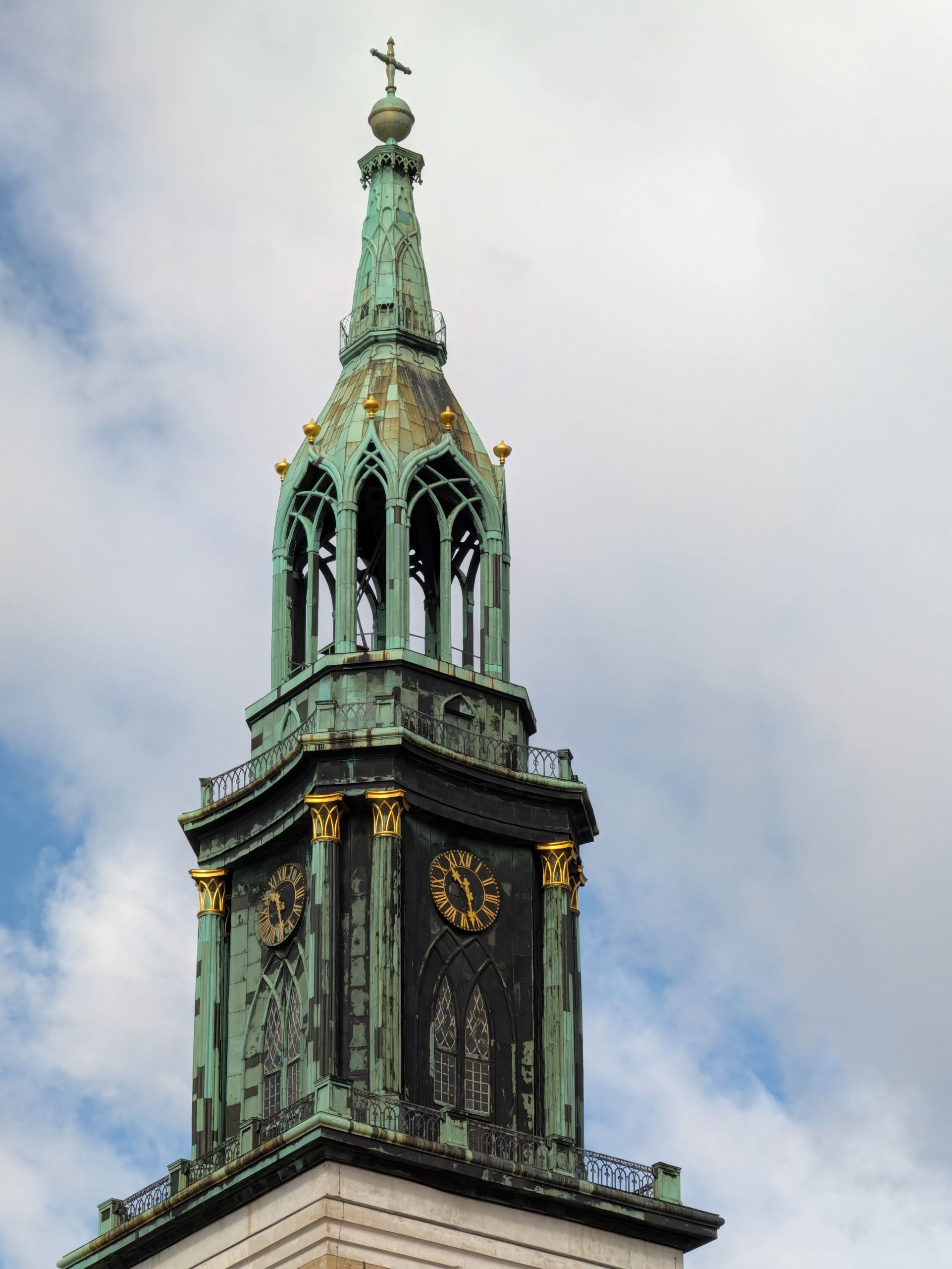 Close-up of a historic green copper church tower with gold clock faces against a cloudy sky.
