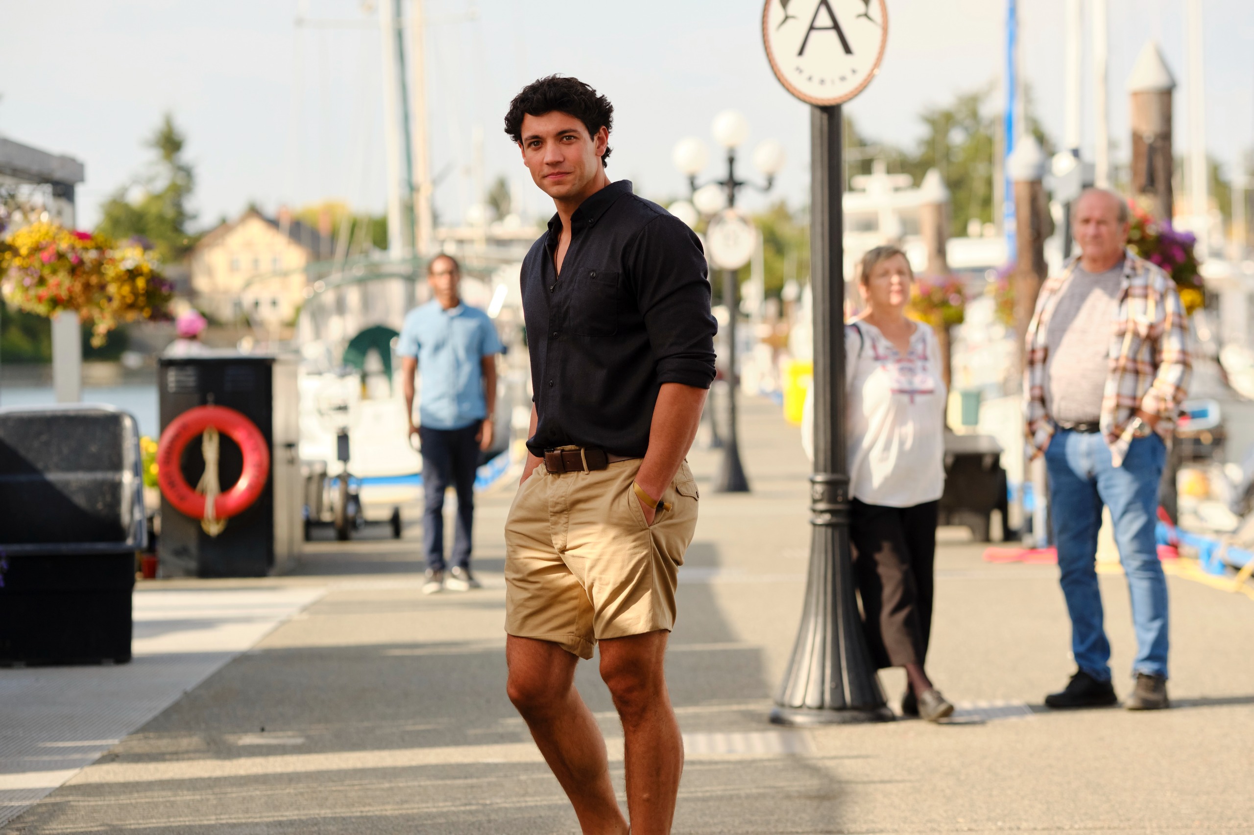 A man in a black polo and khaki shorts (Kyle Clark as Cam Dillinger) stands with his hands in his pockets, on a boardwalk with three people behind him, in Netflix's 'Crew Girl.'