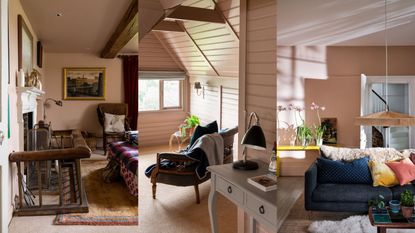 A pink upstairs room with old dark wood beams on the ceiling and wood railings around a stair way / A pink attic room with horizontal painted wood pannelling and a blue chair looking out of a window / A pink living room with a blue couch, yellow side table and a low hanging wooden pendent light 