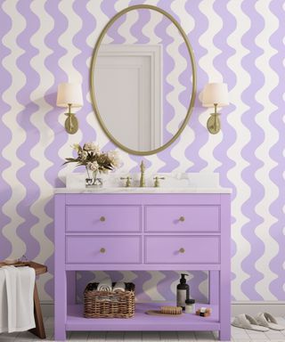 A purple bathroom with white and purple wavy walls, a purple sink unit, and a gold mirror with wall sconces either side