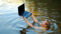 A man submerged in water holding a laptop above the surface.