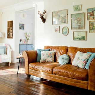 white living room with tan leather sofa, floral cushions, floral vintage paintings on gallery wall, vintage floral armchair, side table, varnished wooden floorboards, blue side console through doorway