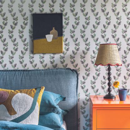 Bedroom with floral wallpaper, bed, orange bedside table and bobbin lamp.