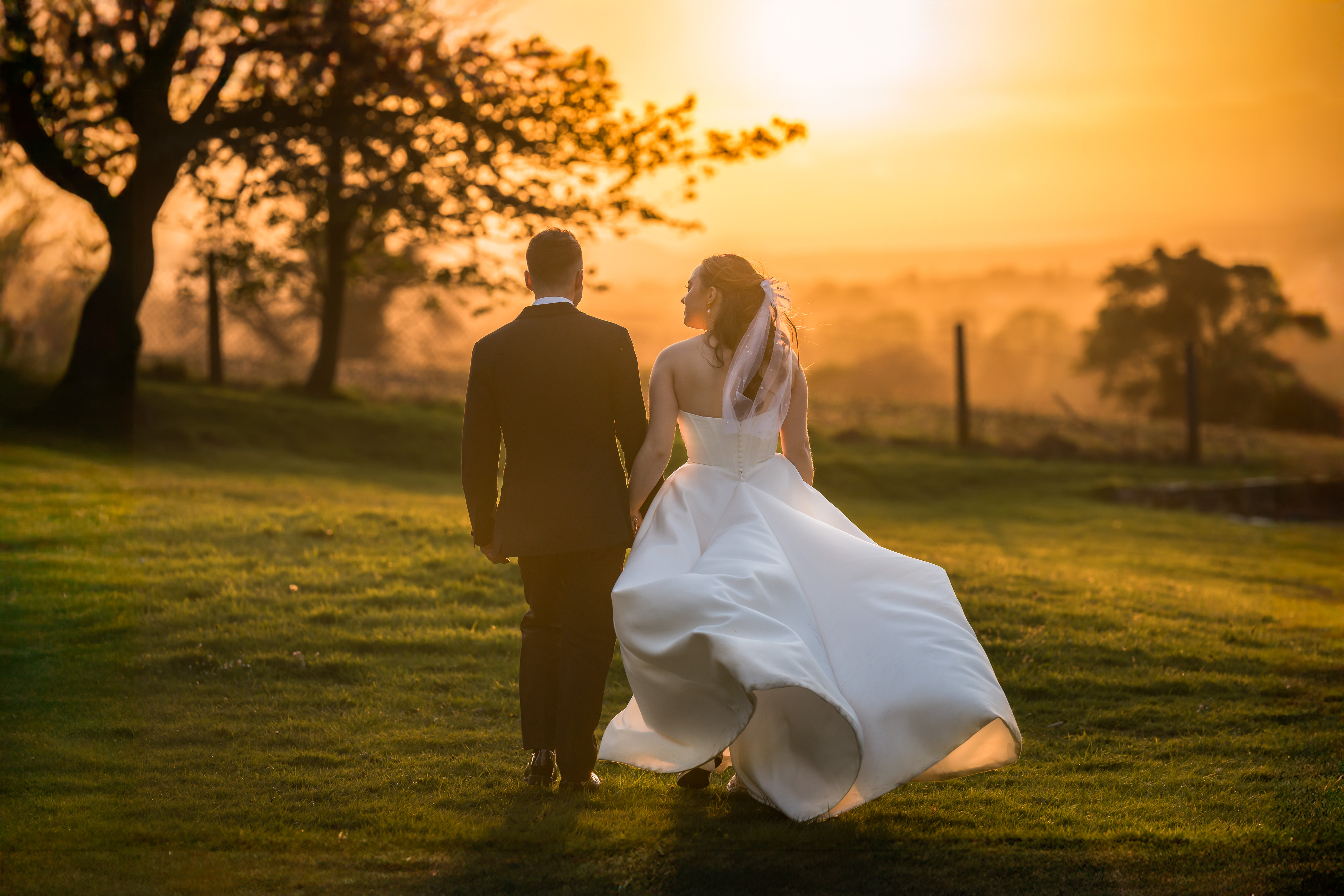 Photograph of a bride and groom in their wedding outfits walking through a field towards a golden sunset. Taken by photographer and educator Nick Church, who will be speaking at The Photography &amp;amp; Video Show 2026