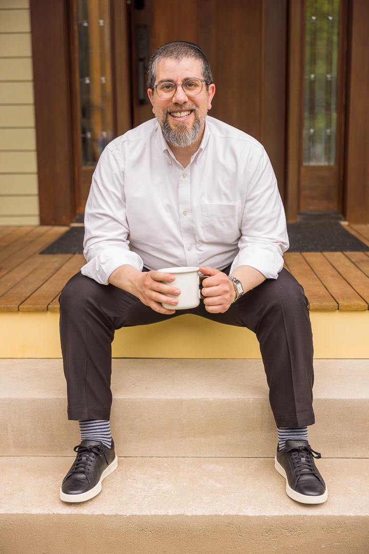 Reuven is sat on a decked front porch, with a coffee mug in his hands. He has a friendly and approachable smile