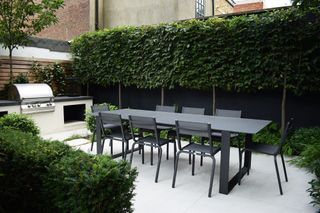 an outdoor kitchen with a dining table surround by privacy hedges