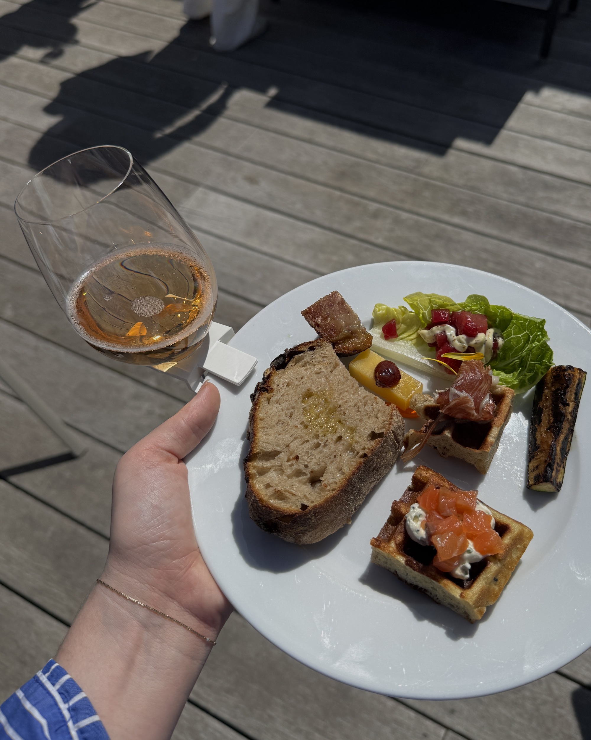 Image of a hand holding a white plate full of different appetizers. A white plate clip holding a wine glass is attached to the plate. The floors below are gray wood.