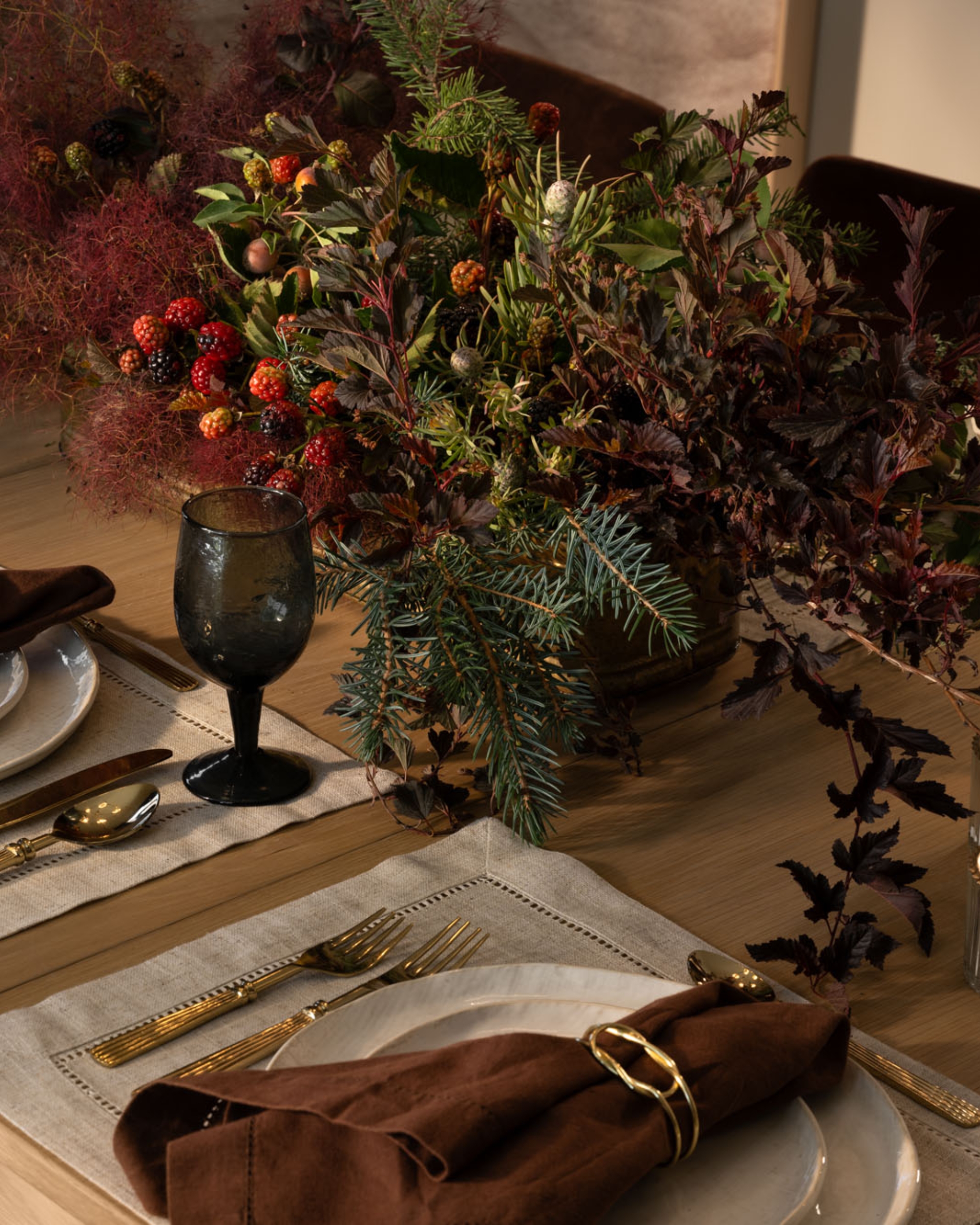 A wooden dining table with a large autumnal foliage centerpiece. There are linen place mats with gold cutlery and brown napkins.
