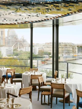 A transitionally decorated restaurant features a layered, mirror ceiling, blue velvety flooring, a wide art wall breaking with the wooden panels stretching across its dining room, and a beautiful, sunlit view over a city.