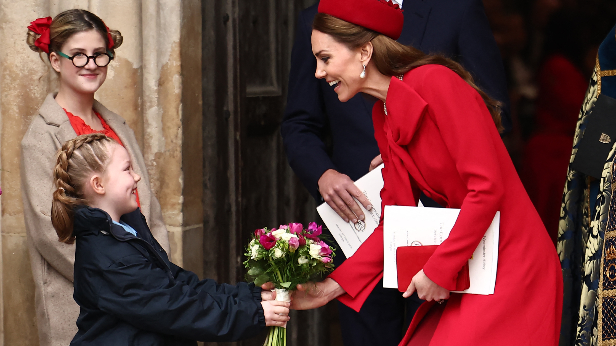 A little girl gives a bouquet to Kate Middleton dressed all in red