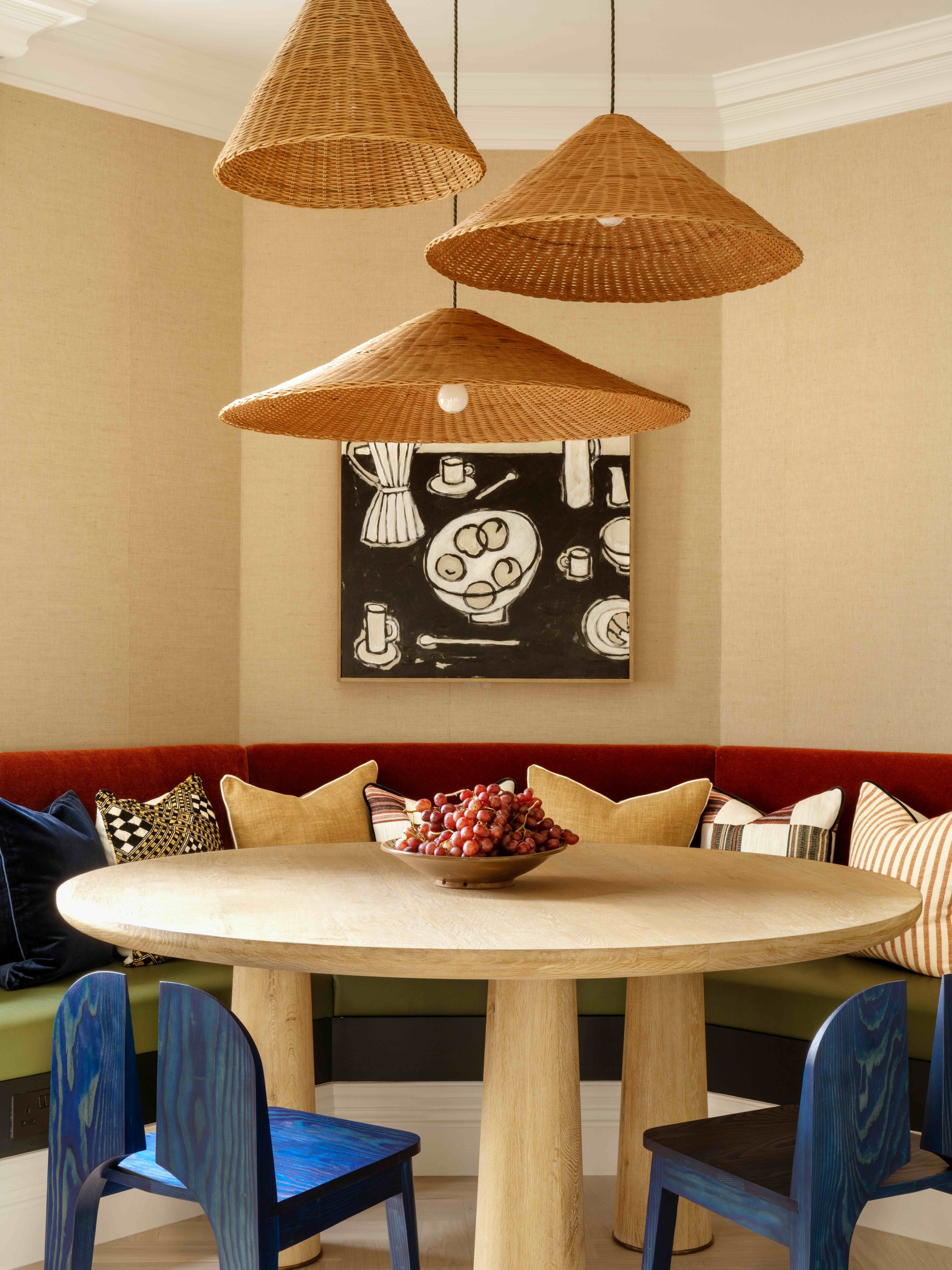 curved corner banquette with red backrest and green seat, lots of cushions, a round timber table, blue dining chairs, and three rattan pendants hanging above