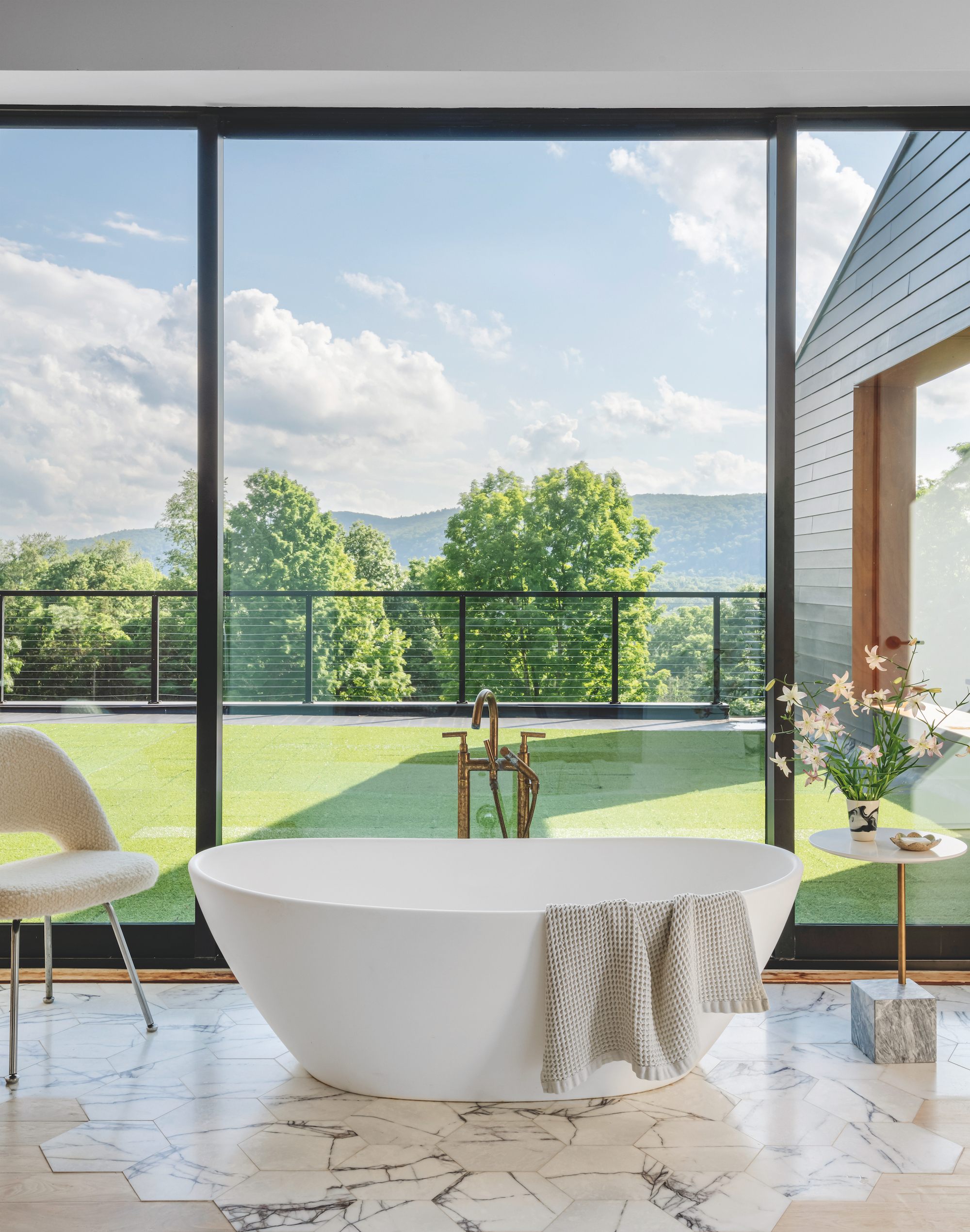 A modern bathroom with floor to ceiling windows with a view of the garden. In front of the windows is a free-standing bathtub, with marble hexagon tiles on the floor and an chair to the left.