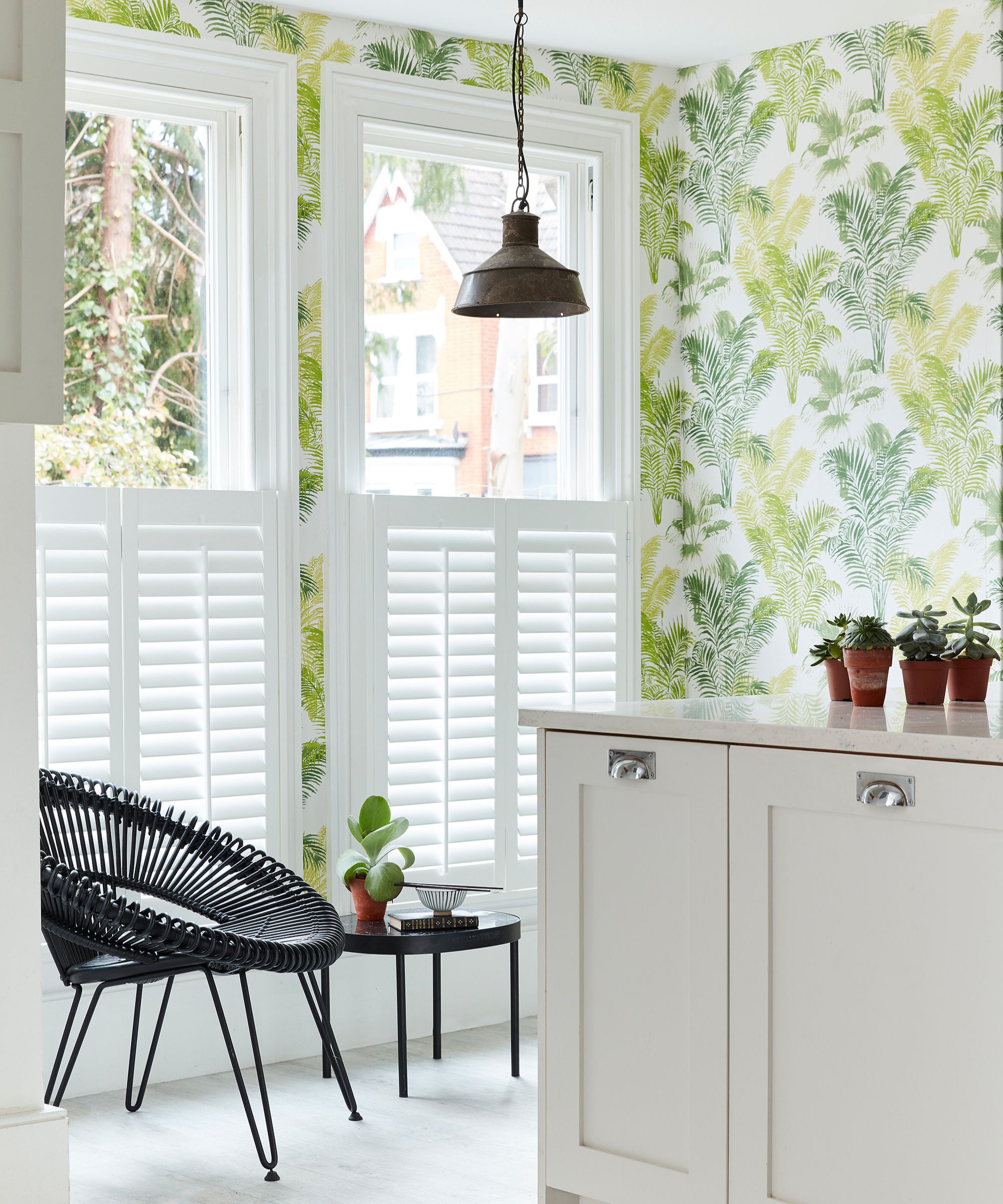 A bright corner room with white cafe-style shutters and palm leaf wallpaper in shades of green and yellow. A black woven chair and small side table with potted plants add contrast.