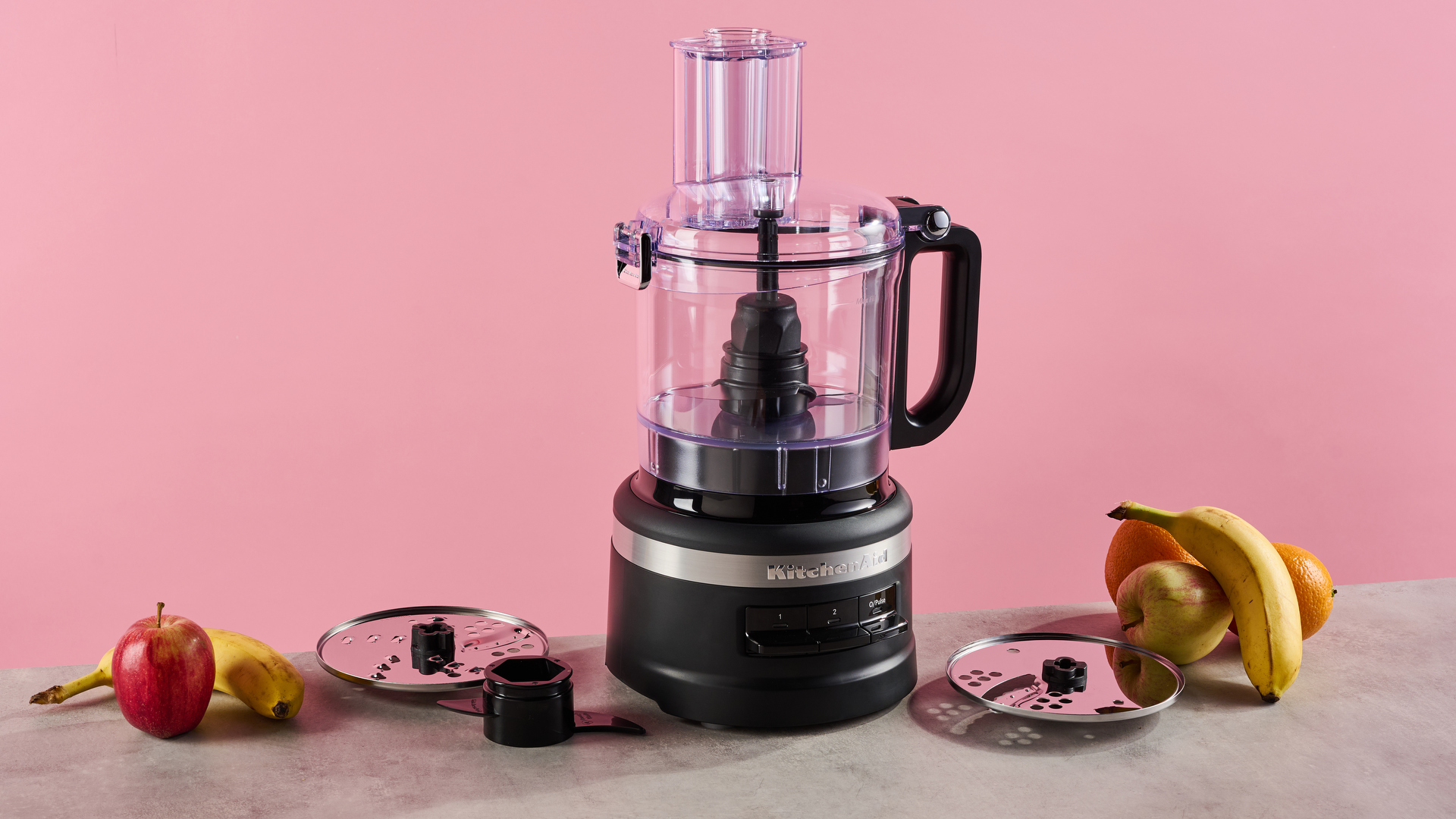 A matte black KitchenAid 7 cup food processor is pictured against a pink background. It is sitting on a grey stone-effect surface on which also sites the reversible slicing and grating disc on the left, with the plastic dough hook positioned just in front, and to the right, the thick slice disc that is only available with the KFP0719 configuration.