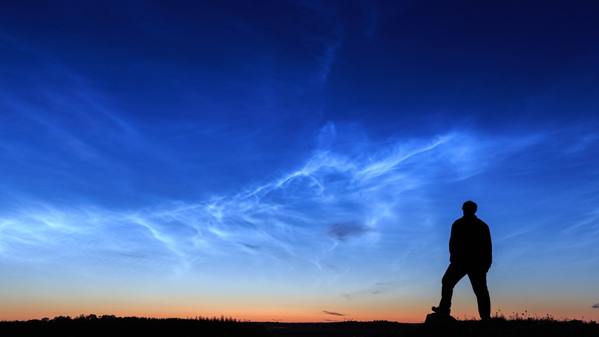 Silhouette of person against dark blue sky with wispy white noctilucent clouds.