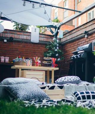 An outdoor courtyard area with a brick wall around it, white and black throw pillows and blankets on the floor, a light wooden table with colorful jars on it, and a black barbecue to the right