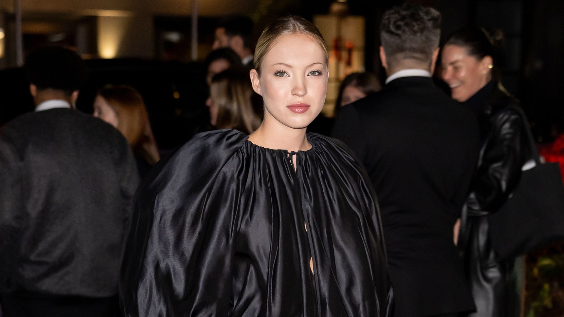 Lila Moss walks a red carpet wearing a black satin balloon blouse with a black miniskirt and black pointed-toe heels.