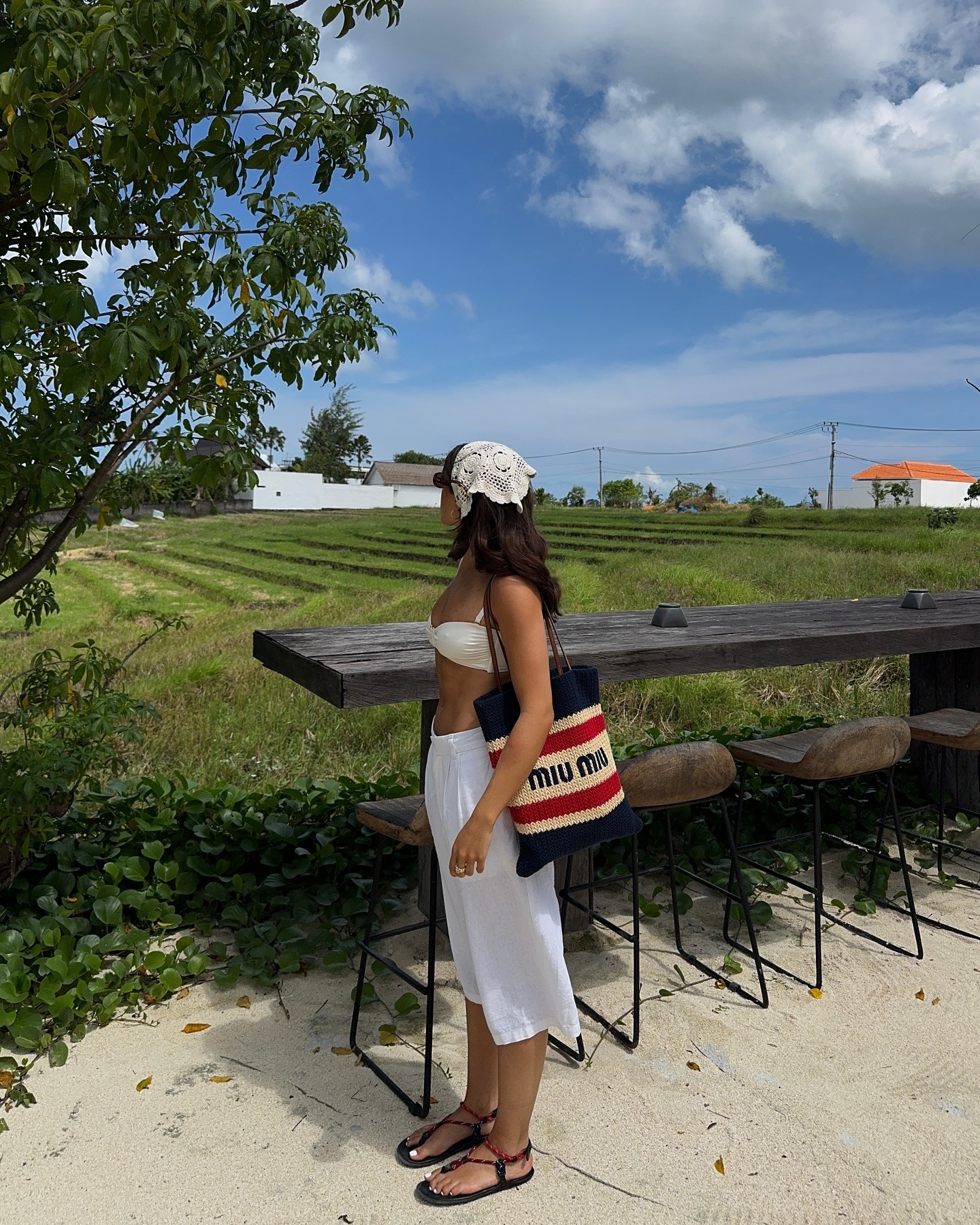 Woman wears bandana, bikini top, long shorts and carries crochet Miu Miu tote bag