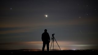 a silhouette of a person standing next to a camera and looking at two bright points of light in the sky, the larger brighter one is Jupiter and the smaller redder one is Mars. A long Perseid meteor streaks through the sky to the right.