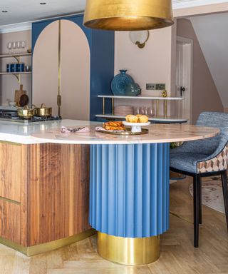A kitchen with pale pink and blue walls, a curved kitchen island with a blue and gold base and wooden cabinets, and a curved gold pendant light hanging above it