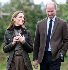 Kate Middleton wearing a green coat and brown cardigan walking next to Prince William in an orchard