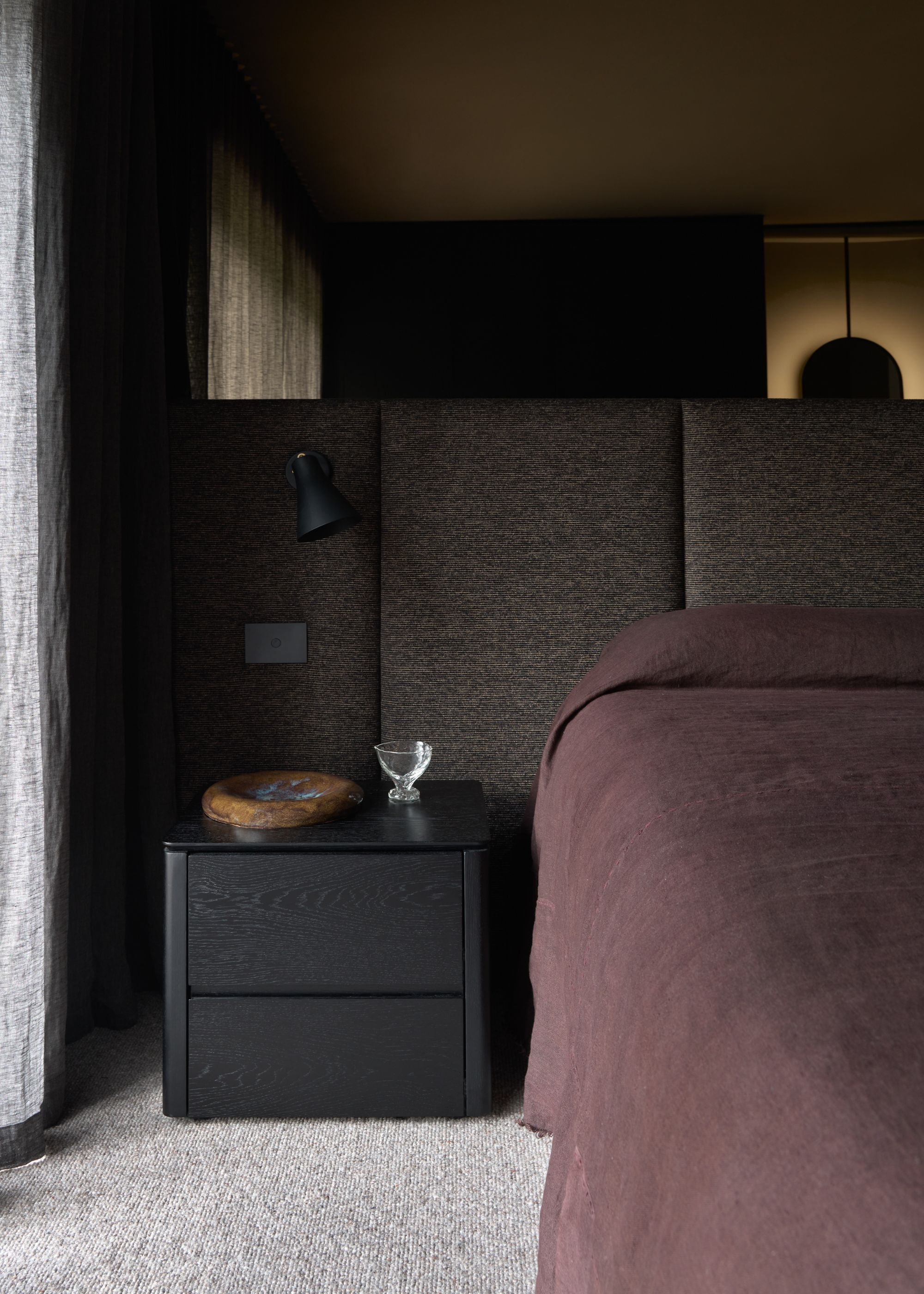 A dark bedroom with an upholstered dark gray headboard, a deep plum bedscape, and a black nightstand with a bowl and a glass