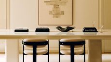 Close-up image of a pale, butter yellow painted office space. There is a long glossy table that has stacks of black books, a black decorative bowl, and two beige and black accent chairs in front of it.