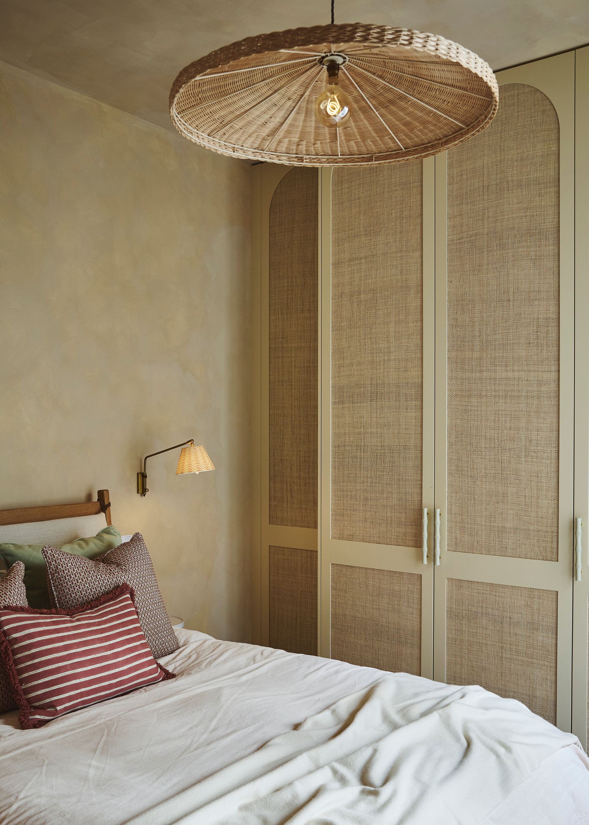 A modern bedroom with sand-colored limewash walls and ceiling, built-in wardrobes with woven front panels, neutral linen bedding with dark red and taupe cushions, and a woven pendant light.