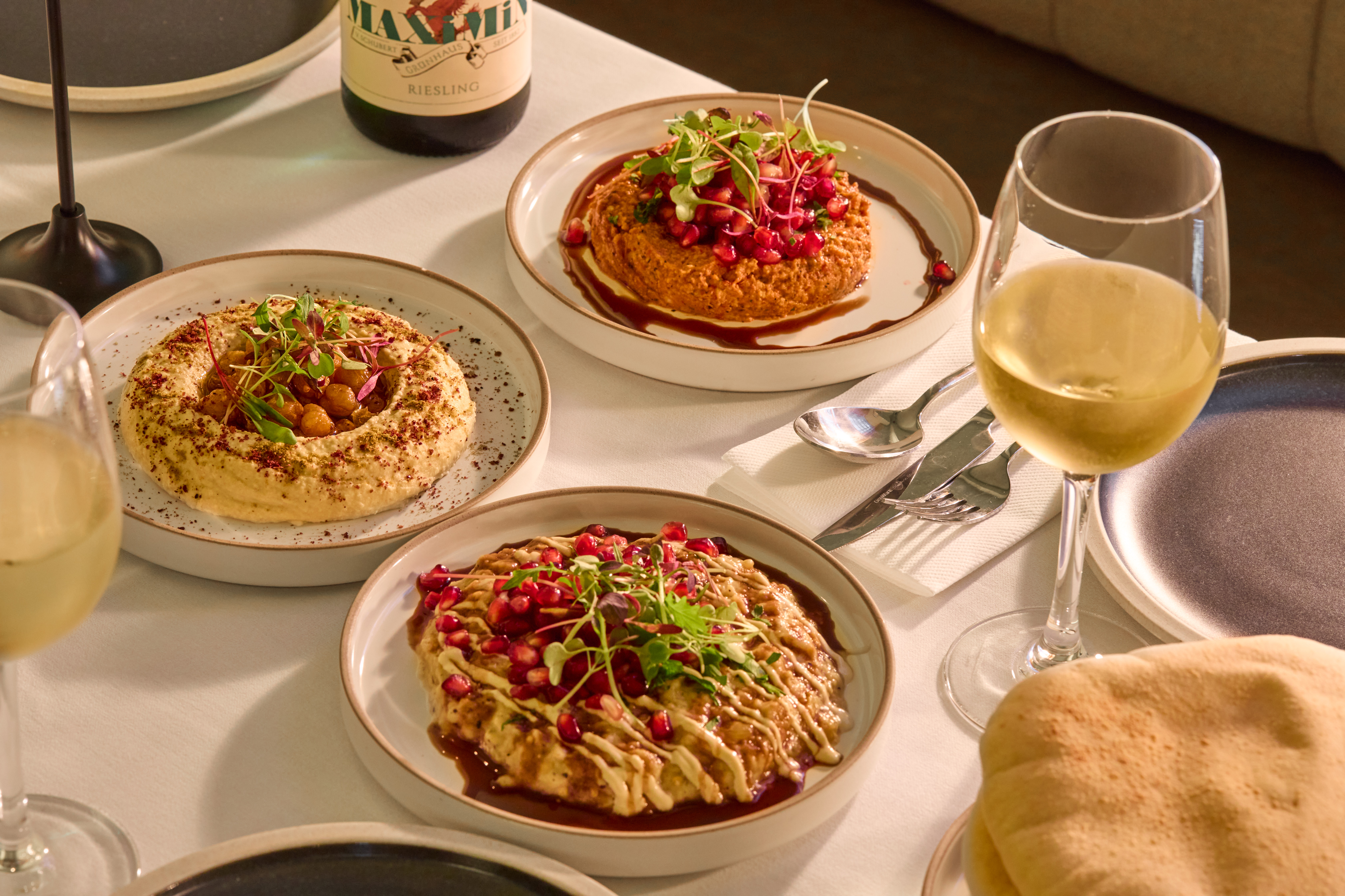 A series of beautifully plated dishes topped with radishes, pomegranate seeds, and spices and placed atop a white table cloth-topped table alongside ceramic plates, bread baskets, and white wine glasses.