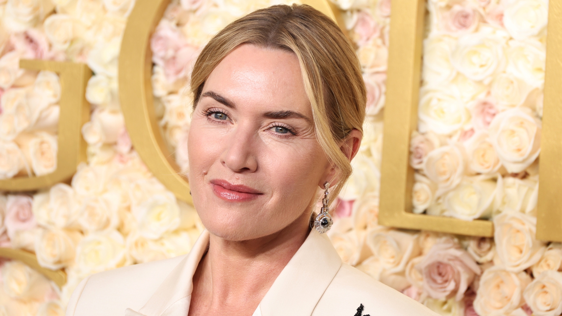 Kate Winslet smiling with her blonde hair in an up-do and wearing a cream blazer