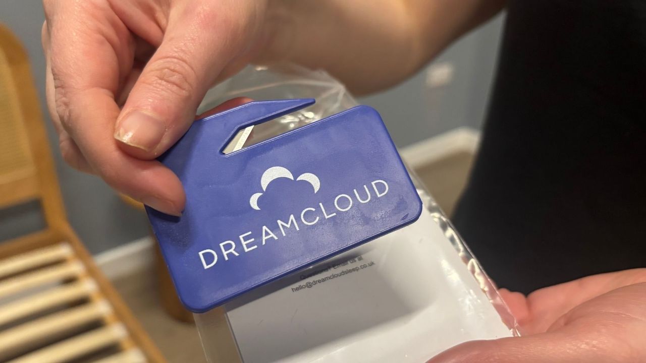 Two mattress testes unbox the DreamCloud Luxe Memory Foam mattress, taking it out of its box, cutting it out of its plastic wrapping and placing it on a white bed frame