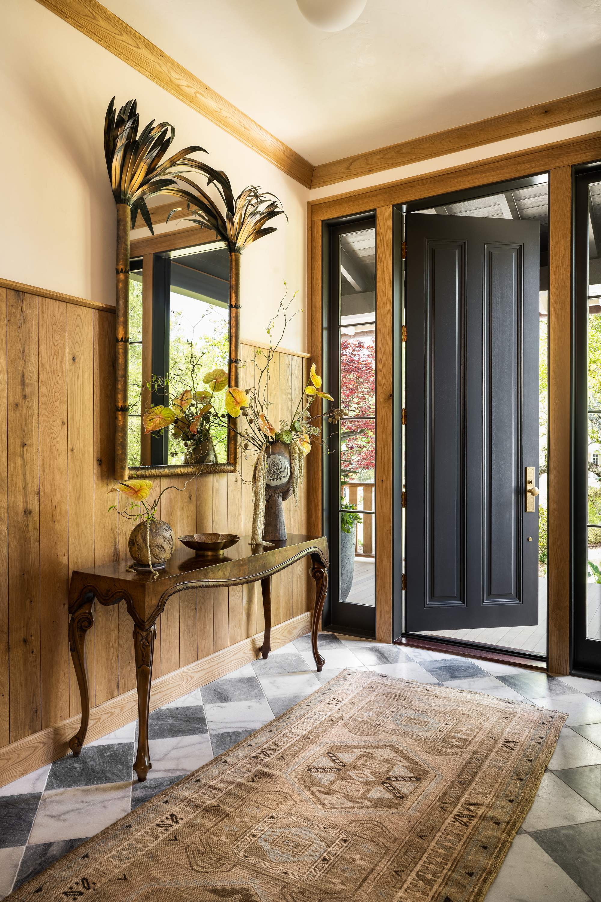 hallway with checkered floors, black front door and timber trim, antique runner, console table with vases of flowers, and gold mirror with feathers