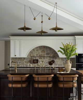 A kitchen with a dark wood island, brown leather counter stools, white cabinetry, and an exposed brick backsplash decorated with a brass pot rail