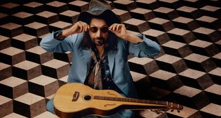 A portrait of Mike Campbell wearing a wide-brimmed hat and dark sunglasses, as the former Heartbreaker posses with a guitar.