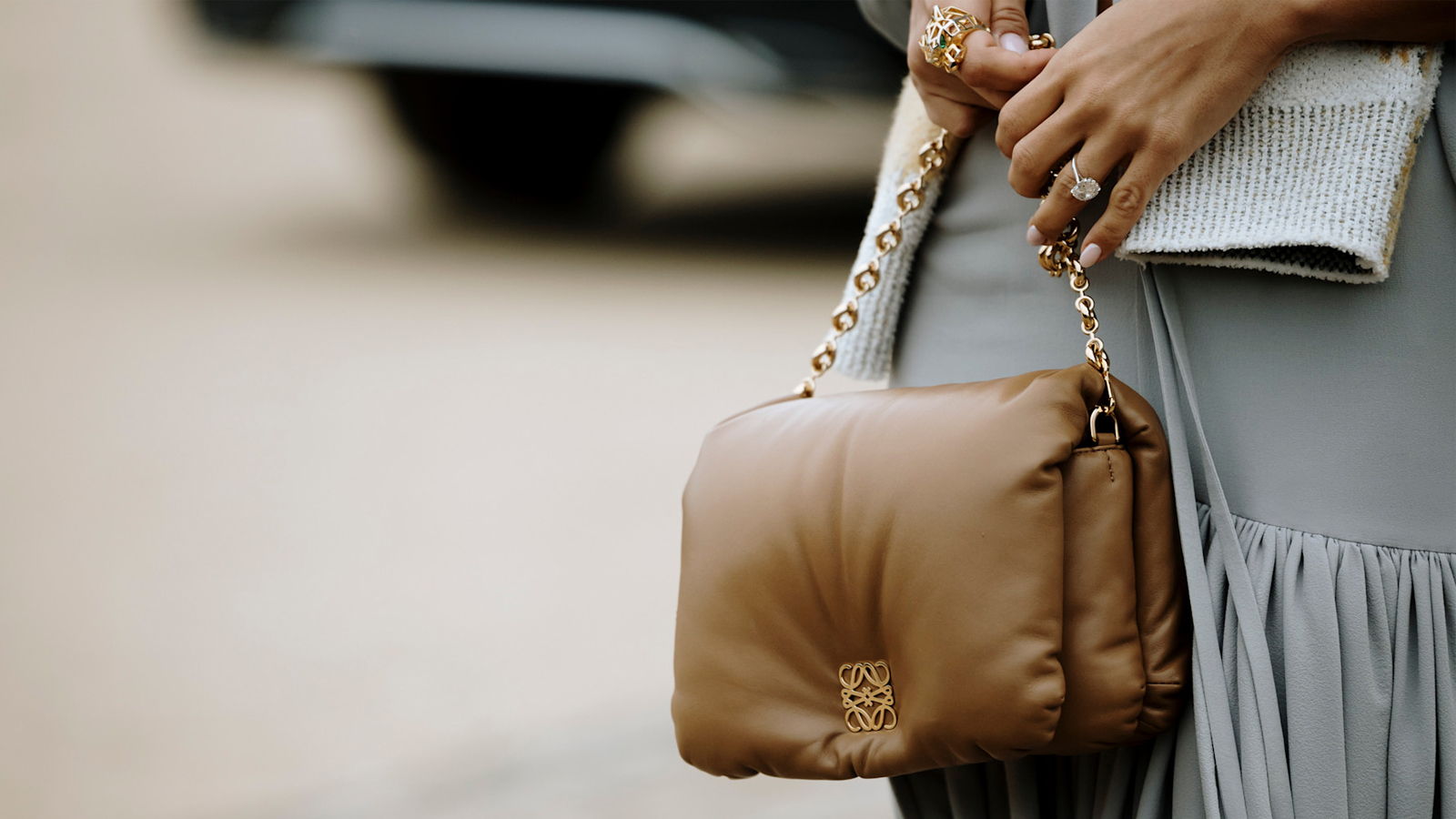 A close up photograph of a woman&#039;s hands, the woman wears an elegant diamond ring, carries a Loewe cloud bag and has an oval shaped manicure with light pink nail polish.