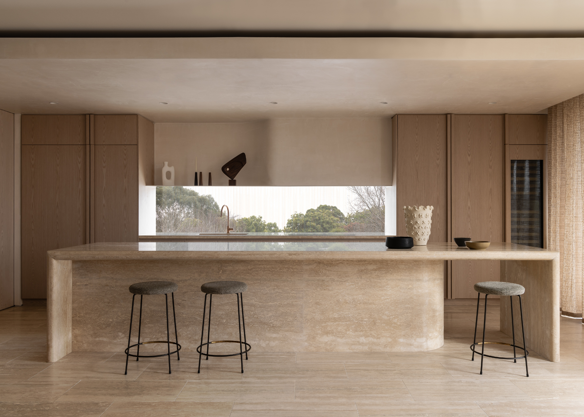 A bright beige minimalist kitchen with a marble island, wood cabinetry, black stools, a white textured vase, and a couple of bowls