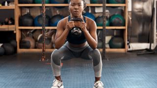 A woman performs a kettlebell goblet squat in a gym. She holds the kettlebell with both hands close to her chest and bends her knees and hips to move close to the ground. Behind her we see stacked exercise balls.