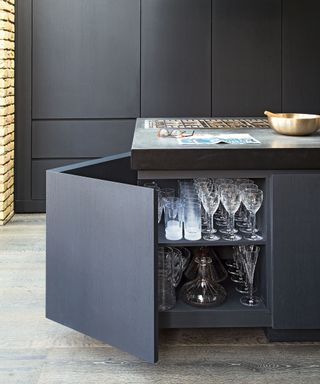 Black kitchen cabinets behind a matching black kitchen island with an open cabinet door showing glasses