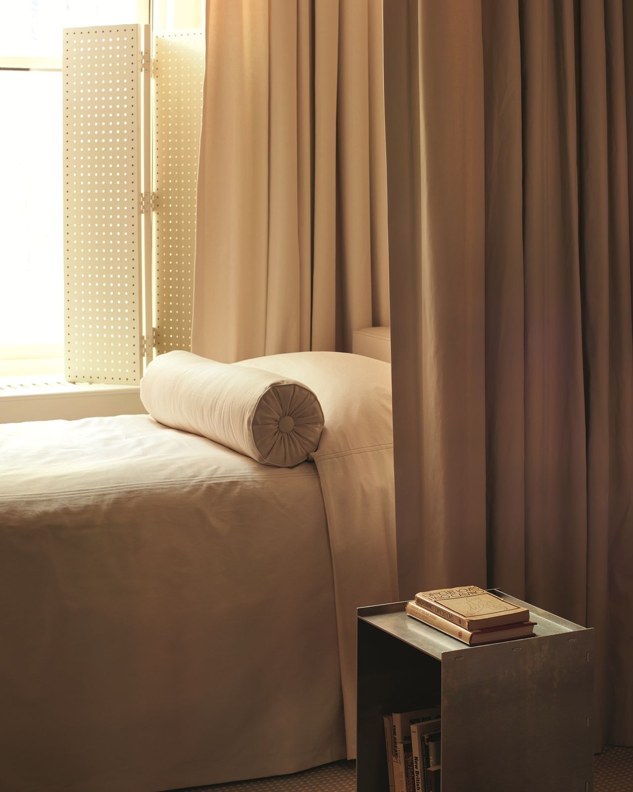 bedroom with curtain behind the bed, metal side table with books, a bolster cushion on bed, and then perforated shutters