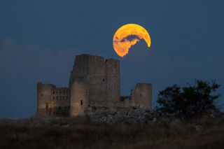 bright golden full moon partially obscured by clouds above a ruin of a large castle.