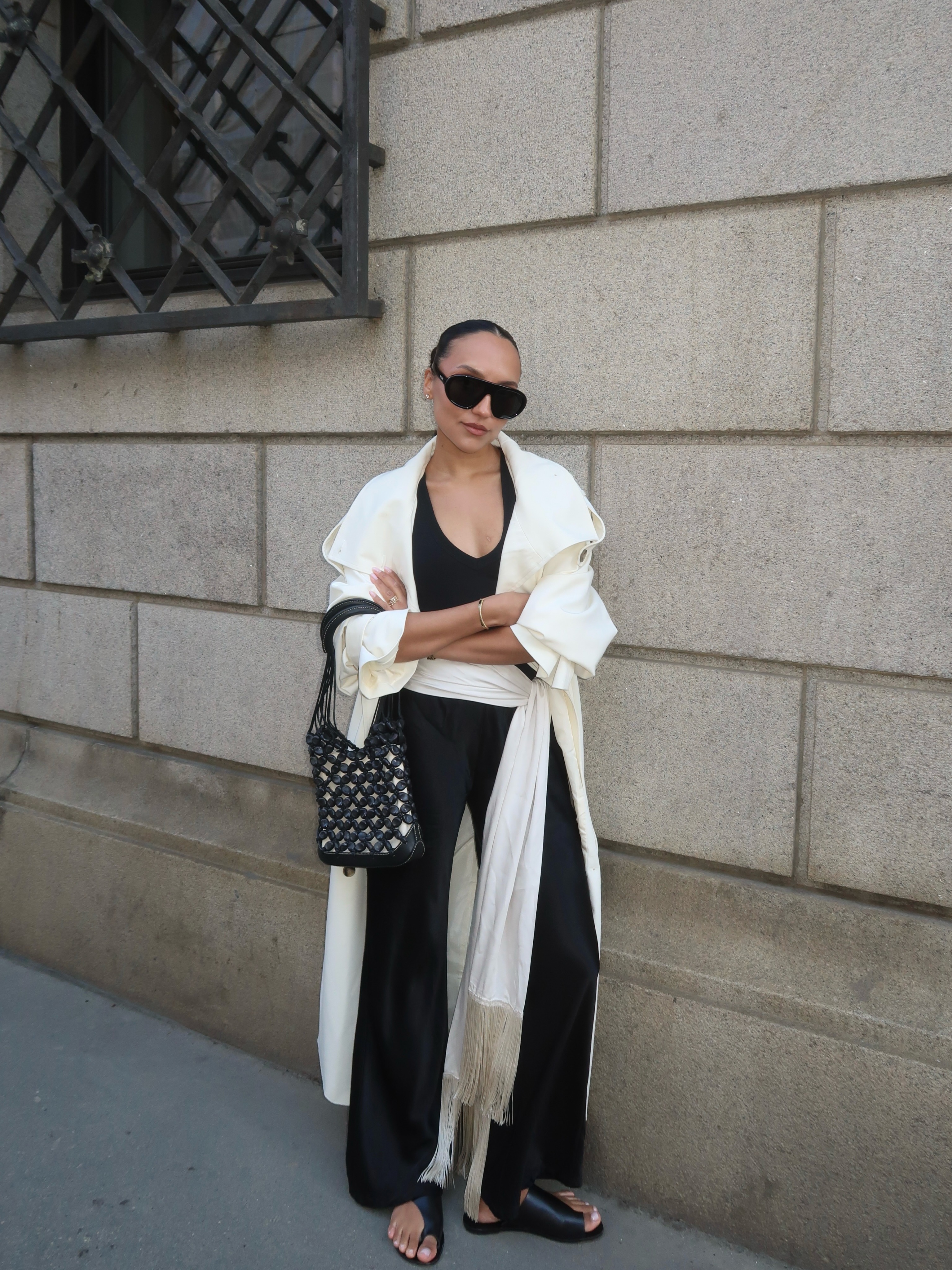 Fashion person Ingrid Edvinsson wears June Outfit Ideas 2026. She wears. A white trench coat, black silk vest, black silk trousers and toe-loop black sandals. She completed the look with a tassel belt.