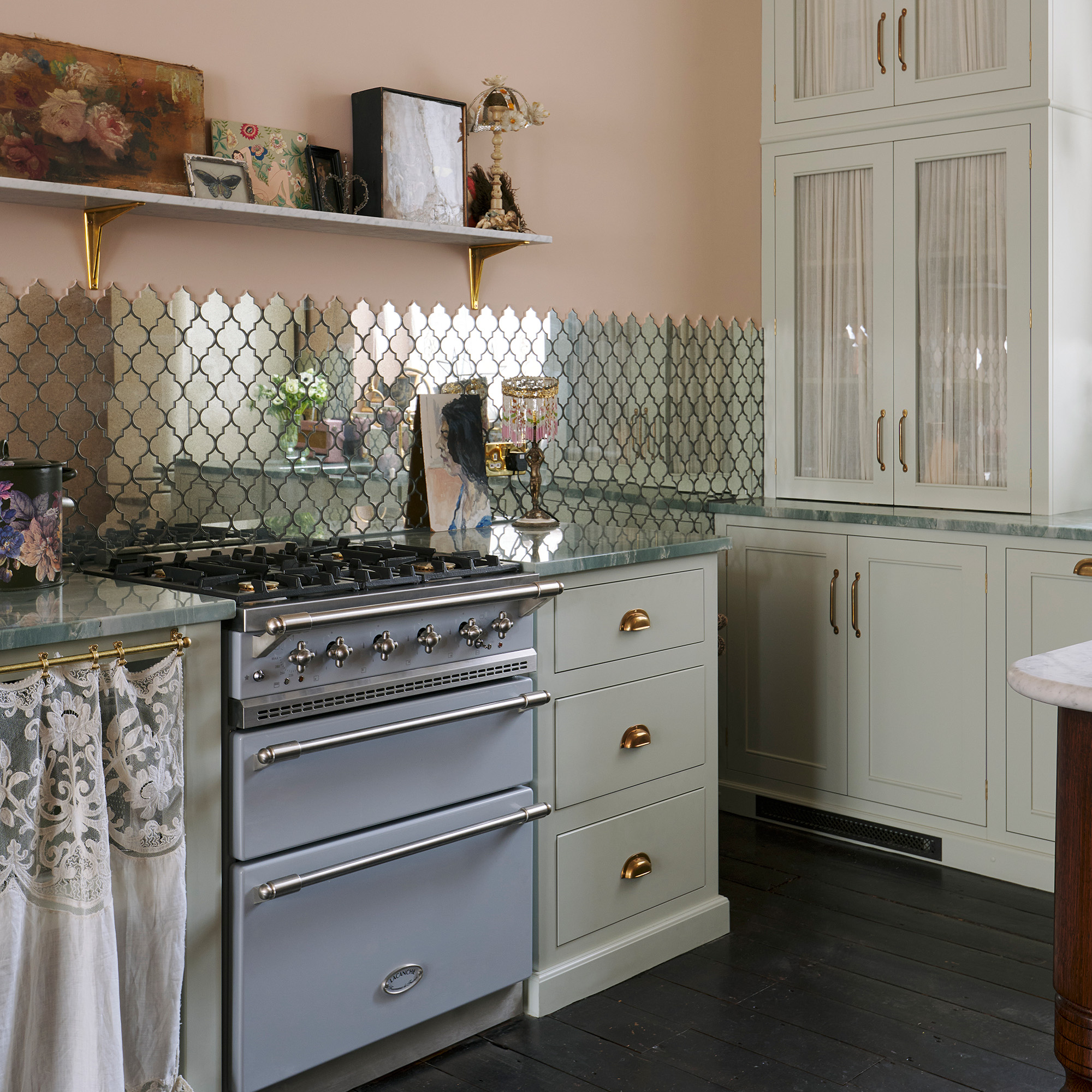 blue range cooker in kitchen with pink walls and dark wood floors cream cabinetry and mirrored tiled splashback with marble open shelf decorated with vintage decor devol