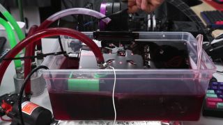 A screenshot of a TrashBench YouTube video, showing a graphics card being lifted out of tub full of transmission fluid used for immersion cooling.